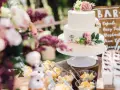 Elegancki tort weselny boho ozdobiony kwiatami, obok deserki i owoce.