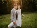 Szczęśliwa para młoda spaceruje po łące. Panna młoda w białej sukni i wianku śmieje się, pan młody w garniturze trzyma ją za rękę. Inspiracja z poradnika weselnego.