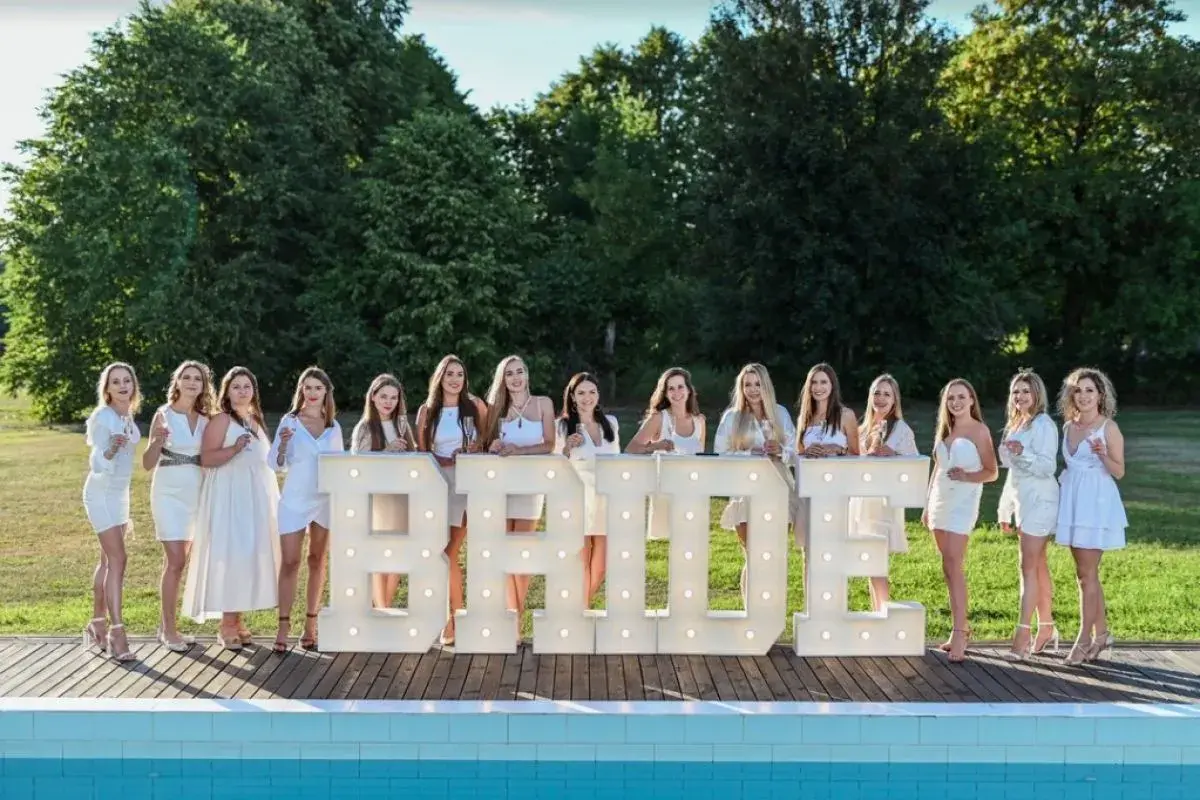 Dziewczyny w białych sukienkach świętują wieczór panieński w plenerze, pozując przy podświetlanych literach "BRIDE".