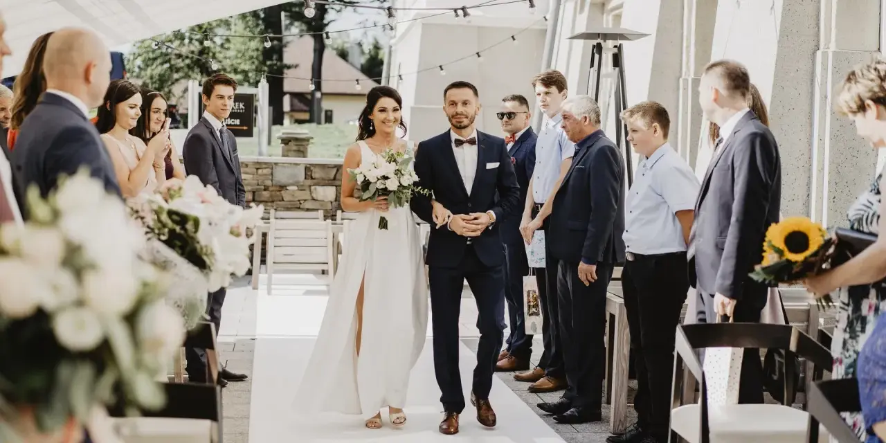 Para młoda idzie do ołtarza na ślubie cywilnym. Panna młoda w białej sukni z bukietem kwiatów, pan młody w granatowym garniturze.