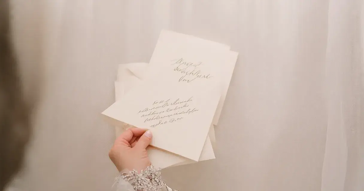 Dłoń w koronkowej sukni trzyma zaproszenia ślubne. Eleganckie zawiadomienia z kaligrafią to piękne zyczenia na slub.