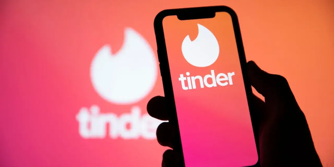 Dłoń trzyma smartfon z logo Tinder. W tle widać większe, rozmyte logo aplikacji, sugerujące nowe tinder powiadomienia.
