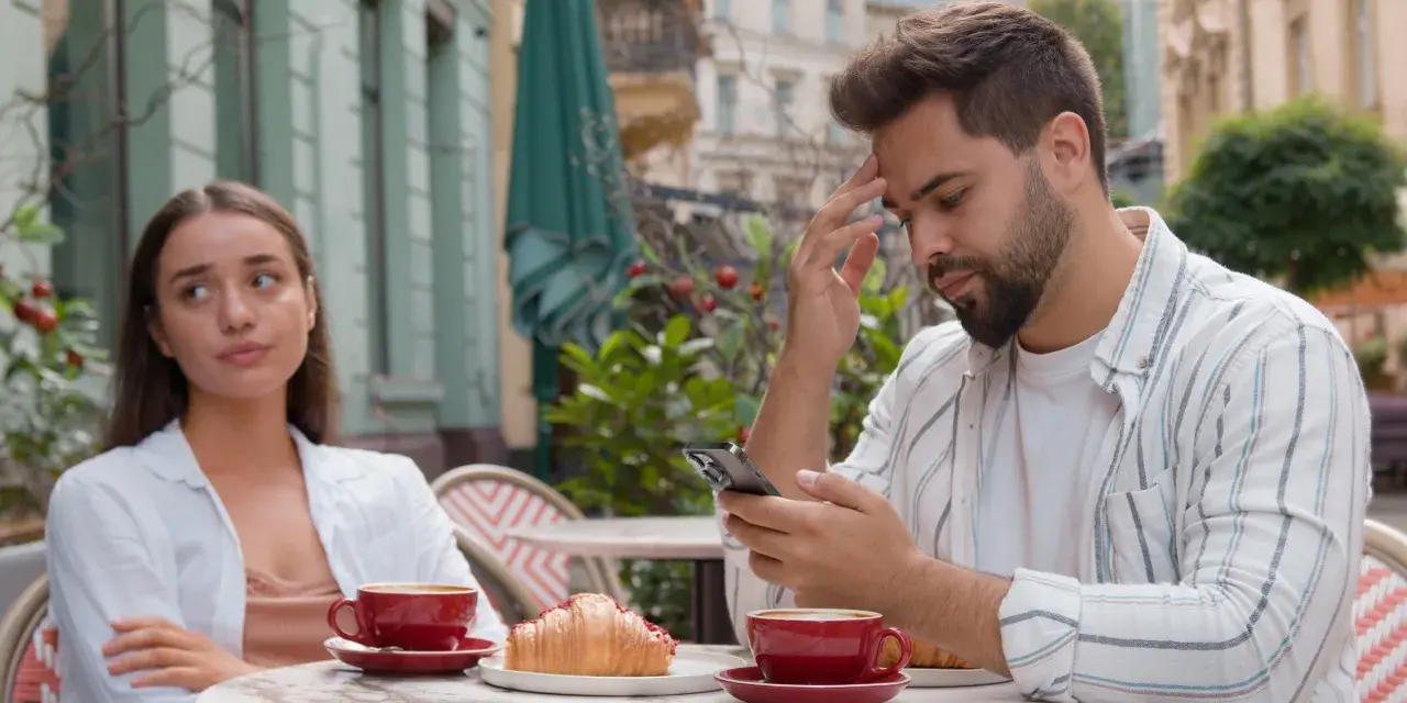 Para siedzi przy stoliku w kawiarni. Mężczyzna zamyślony patrzy w telefon, kobieta obok niego wygląda na znudzoną.