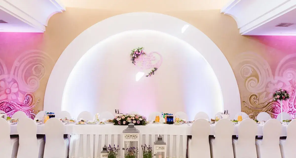 Eleganckie wesele w stylu nowoczesnym. Długi stół weselny nakryty białym obrusem, ozdobiony kwiatami i lampionami. W tle serce z imionami pary młodej.