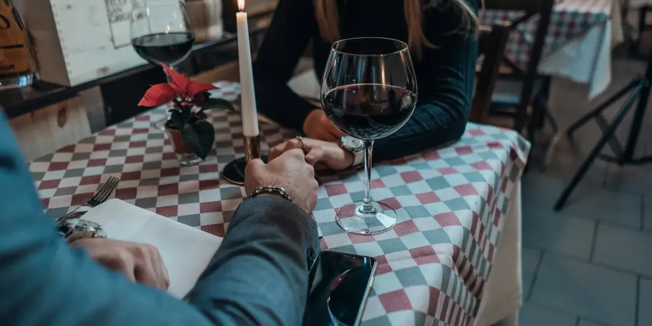 Romantyczna kolacja, świeca, wino i dłonie splecione na stole. Widać, jak angażuje się mężczyzna, skupiony na partnerce.