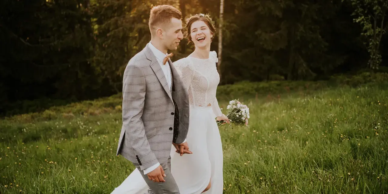 Szczęśliwa para młoda spaceruje po łące. Panna młoda w białej sukni i wianku śmieje się, pan młody w garniturze trzyma ją za rękę. Inspiracja z poradnika weselnego.
