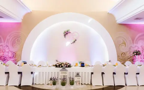 Eleganckie wesele w stylu nowoczesnym. Długi stół weselny nakryty białym obrusem, ozdobiony kwiatami i lampionami. W tle serce z imionami pary młodej.