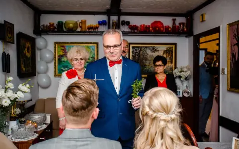 Uroczyste błogosławieństwo młodej pary przez starszego mężczyznę w niebieskiej marynarce.