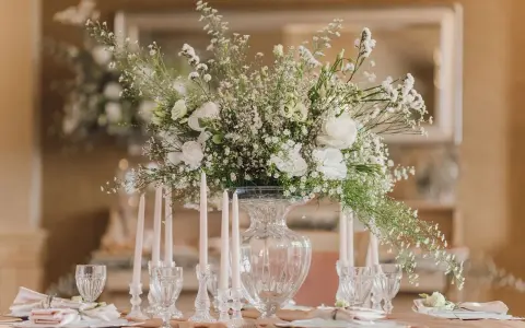 Eleganckie ustawienie stołów na 40 osób. Centralny punkt stanowi bukiet białych kwiatów w szklanym wazonie, otoczony świecami i kryształowymi kieliszkami.