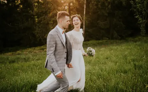 Szczęśliwa para młoda spaceruje po łące. Panna młoda w białej sukni i wianku śmieje się, pan młody w garniturze trzyma ją za rękę. Inspiracja z poradnika weselnego.
