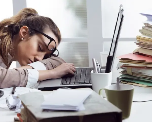 Zmęczona kobieta śpi na laptopie, otoczona stertami dokumentów. To obrazek pokazujący skutki związku z pracoholikiem.
