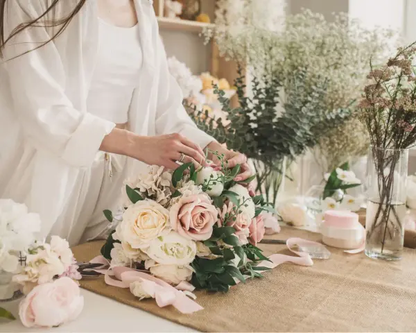 Dłonie florystki tworzą piękny, pastelowy bukiet ślubny. W tle widać inne kwiaty i akcesoria.