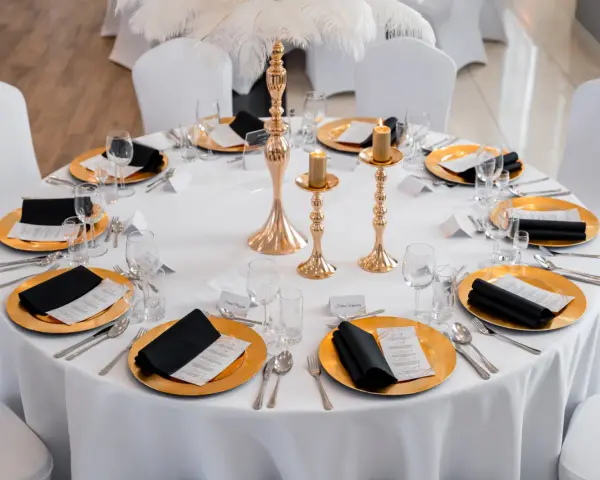 Elegancko nakryty stół weselny ze złotymi talerzami, czarnymi serwetkami i świecami. Wesele motywy złota i bieli.