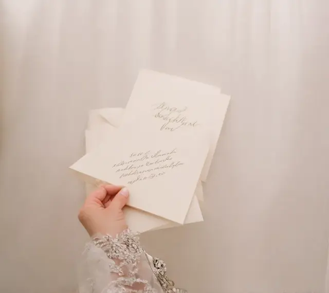 Dłoń w koronkowej sukni trzyma zaproszenia ślubne. Eleganckie zawiadomienia z kaligrafią to piękne zyczenia na slub.