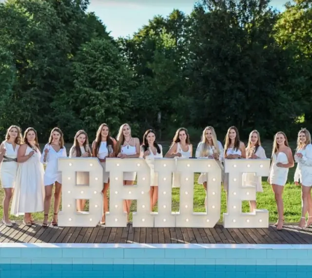 Dziewczyny w białych sukienkach świętują wieczór panieński w plenerze, pozując przy podświetlanych literach "BRIDE".