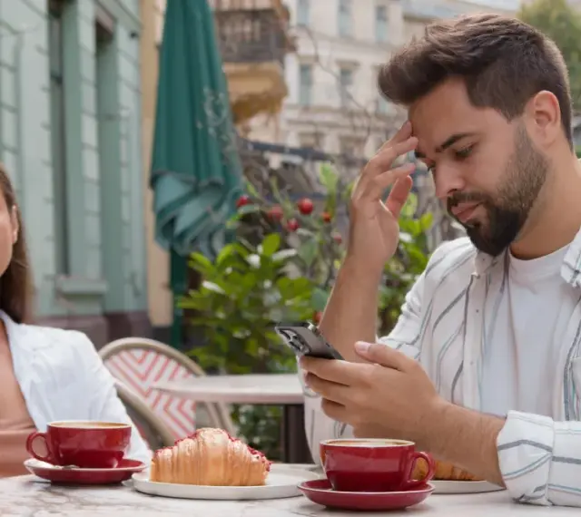 Para siedzi przy stoliku w kawiarni. Mężczyzna zamyślony patrzy w telefon, kobieta obok niego wygląda na znudzoną.