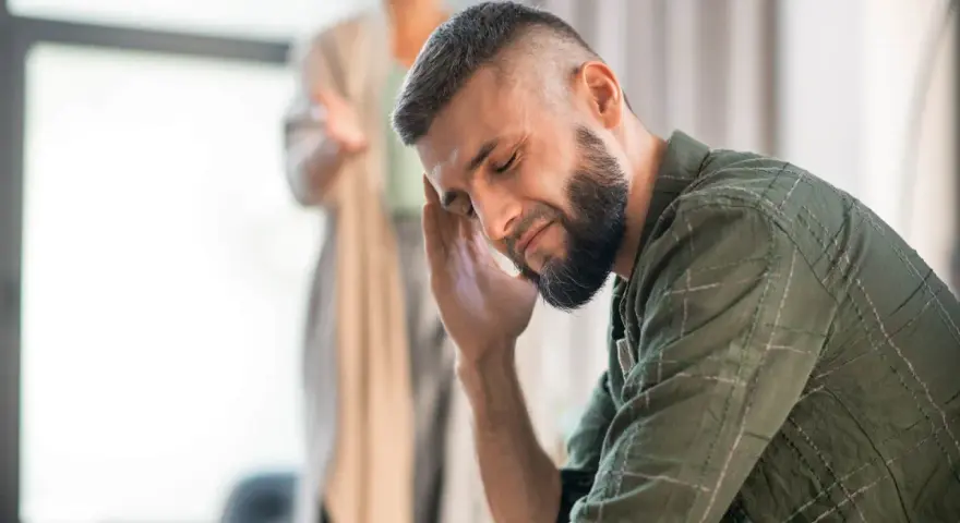 Mężczyzna z ręką na skroni, wyrażający zaangażowanie emocjonalne. W tle niewyraźna postać.