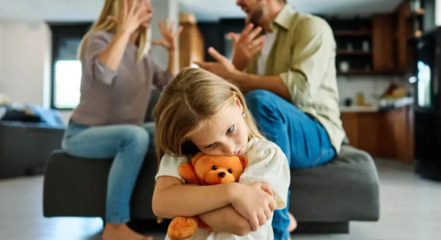 Smutna dziewczynka tuli misia, podczas gdy rodzice kłócą się w tle. To trudny czas, gdy rozwód dotyka dzieci.