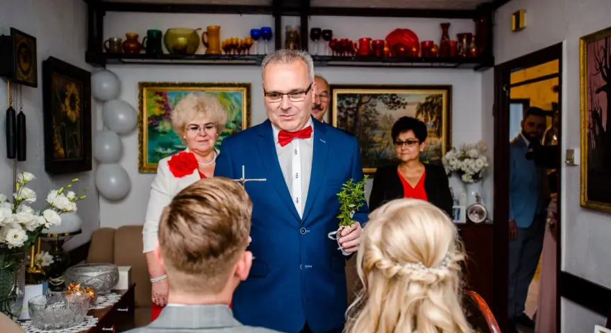 Uroczyste błogosławieństwo młodej pary przez starszego mężczyznę w niebieskiej marynarce.