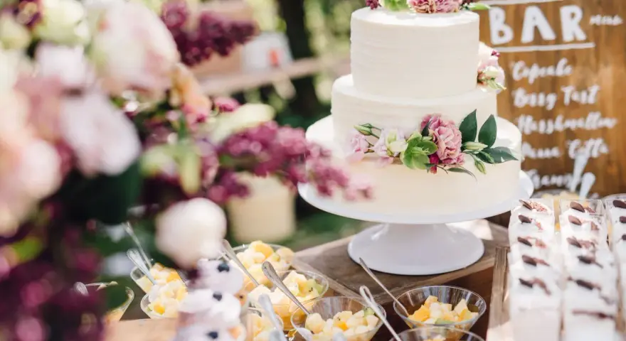 Elegancki tort weselny boho ozdobiony kwiatami, obok deserki i owoce.
