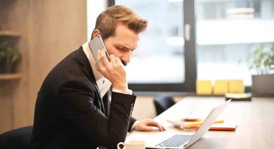 Mężczyzna w garniturze prowadzi ważną rozmowę przez telefon, siedząc przy biurku z laptopem i filiżanką kawy.