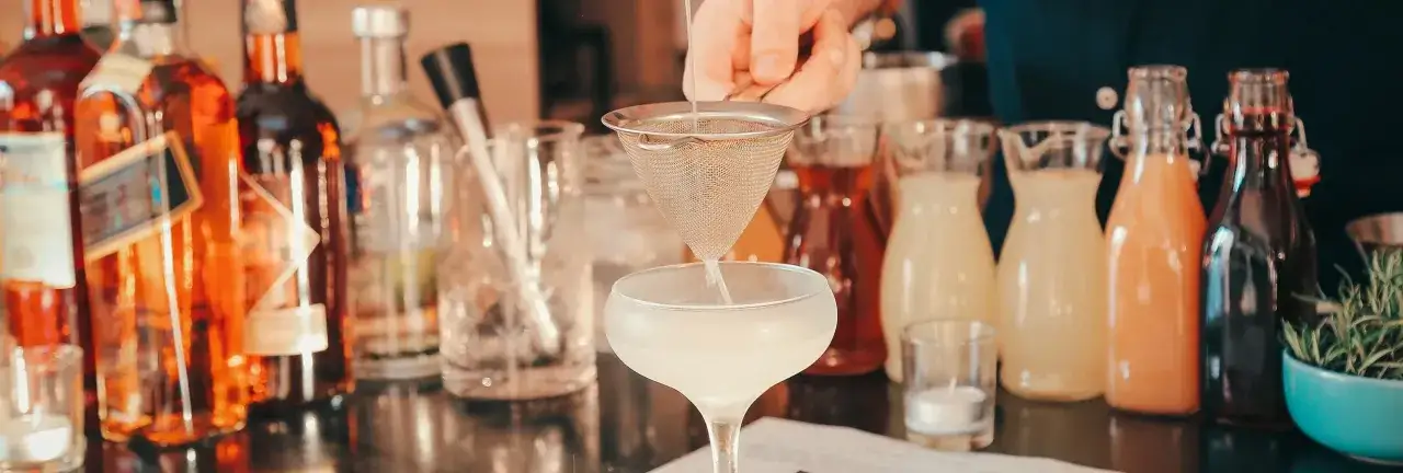 Barman przygotowuje drinki na wesele. Na blacie stoją butelki z alkoholami i syropami, a do kieliszka przesącza się płyn.