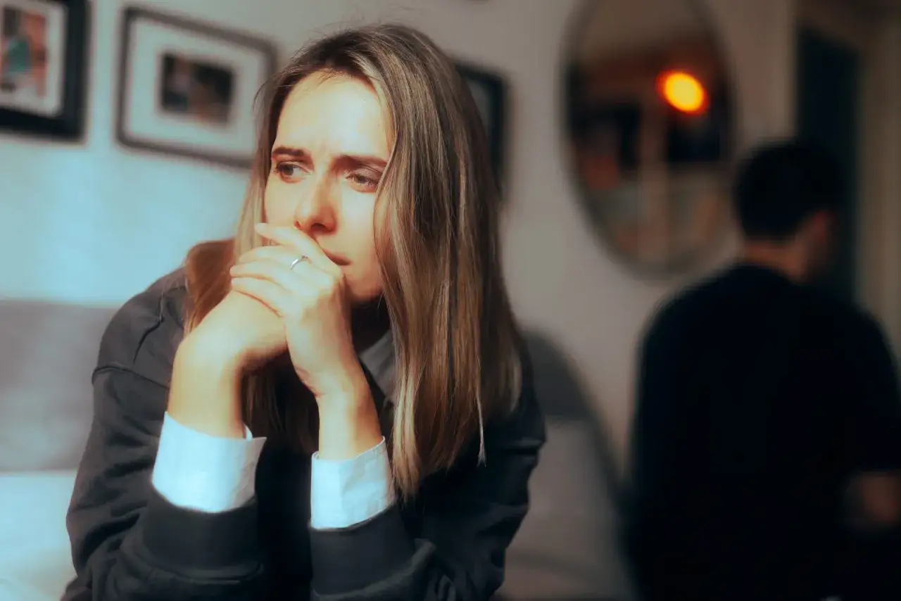 Kobieta zamyślona, z rękami przy twarzy, w tle niewyraźna postać mężczyzny. Widoczny dystans emocjonalny między nimi.