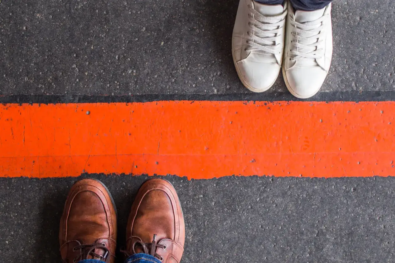 Dwie pary butów, jedna w brązowych, druga w białych sneakersach, stoją po przeciwnych stronach pomarańczowej linii. To symbol stawiania granic facetowi.