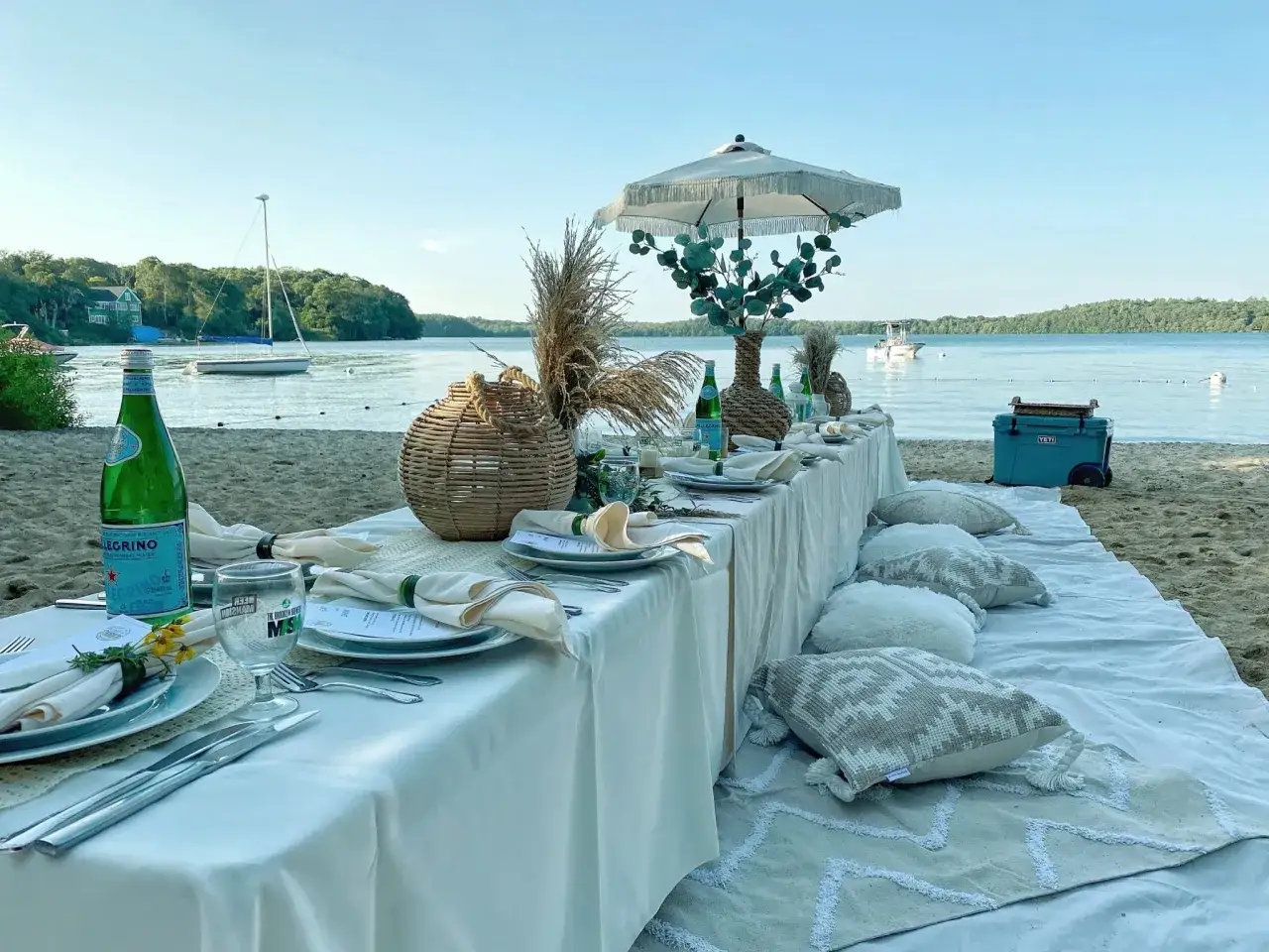 Elegancki wieczór panieński w plenerze nad jeziorem. Stół z nakryciami, parasol plażowy i poduszki tworzą przytulną atmosferę.