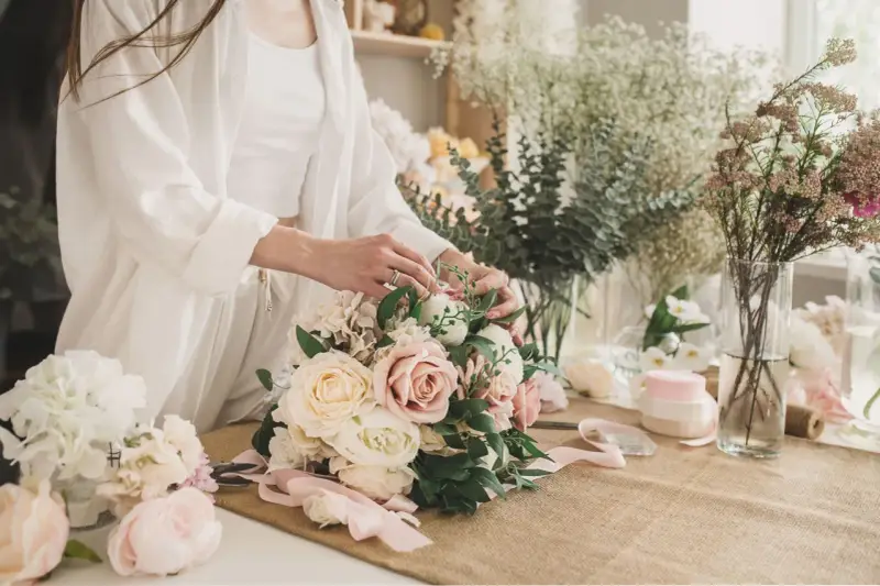 Dłonie florystki tworzą piękny, pastelowy bukiet ślubny. W tle widać inne kwiaty i akcesoria.