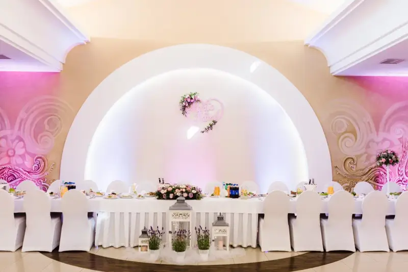 Eleganckie wesele w stylu nowoczesnym. Długi stół weselny nakryty białym obrusem, ozdobiony kwiatami i lampionami. W tle serce z imionami pary młodej.