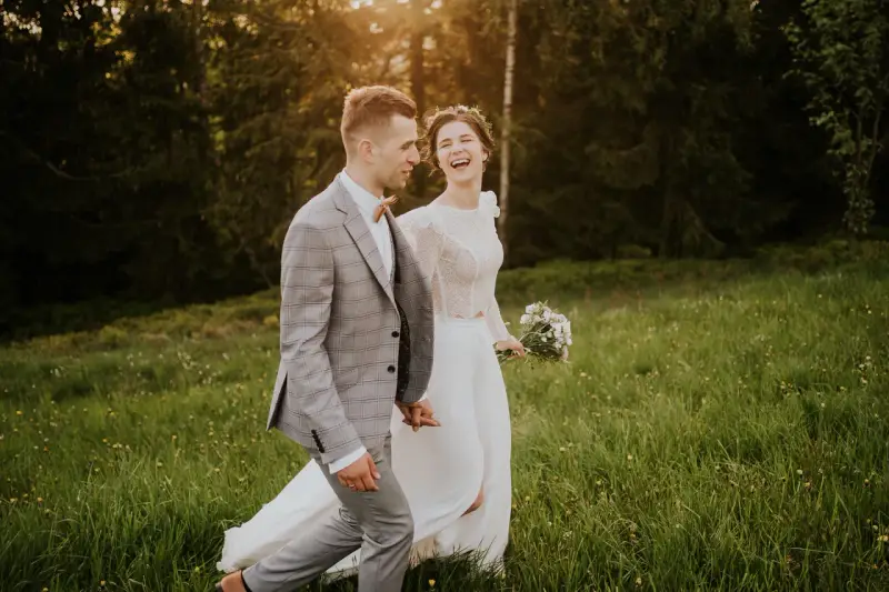 Szczęśliwa para młoda spaceruje po łące. Panna młoda w białej sukni i wianku śmieje się, pan młody w garniturze trzyma ją za rękę. Inspiracja z poradnika weselnego.